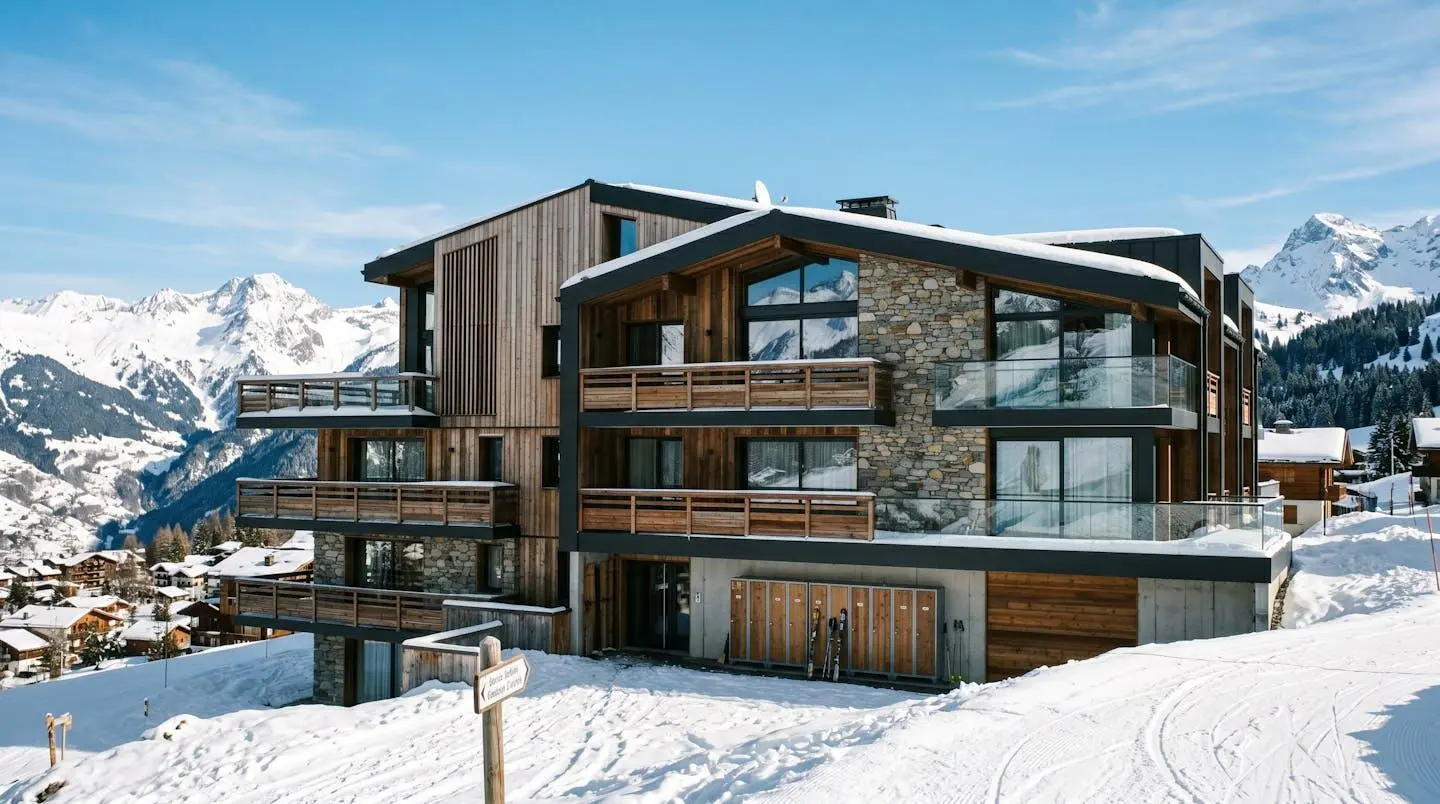Façade d'une résidence de ski contemporaine avec plusieurs balcons orientés sud et casiers à ski visibles au rez-de-chaussée sous un ciel bleu lumineux